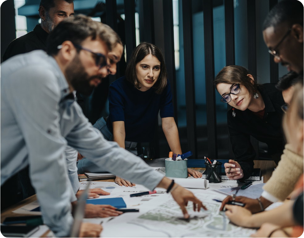Eine Gruppe von Personen versammelt sich um einen großen Tisch, diskutiert engagiert und zeigt auf Unterlagen und Diagramme. Die Atmosphäre ist konzentriert und kooperativ, mehrere Personen beugen sich vor, um an der Diskussion teilzunehmen. Auf dem Tisch liegen diverse Büromaterialien. A group of people gathered around a large table, actively discussing and pointing at documents and diagrams. The atmosphere is focused and collaborative, with several individuals leaning in to engage in the discussion. Office supplies are visible on the table.