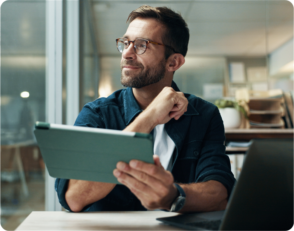 Ein Mann mit Brille und Bart sitzt an einem Schreibtisch in einem modernen Büro, hält ein Tablet in der Hand und blickt nachdenklich zur Seite. Vor ihm steht ein offener Laptop, im Hintergrund sind Regale mit Dokumenten zu sehen. Man with glasses and a beard sitting at a desk in a modern office, holding a tablet and looking thoughtfully to the side. A laptop is open in front of him, and office shelves with documents are visible in the background.